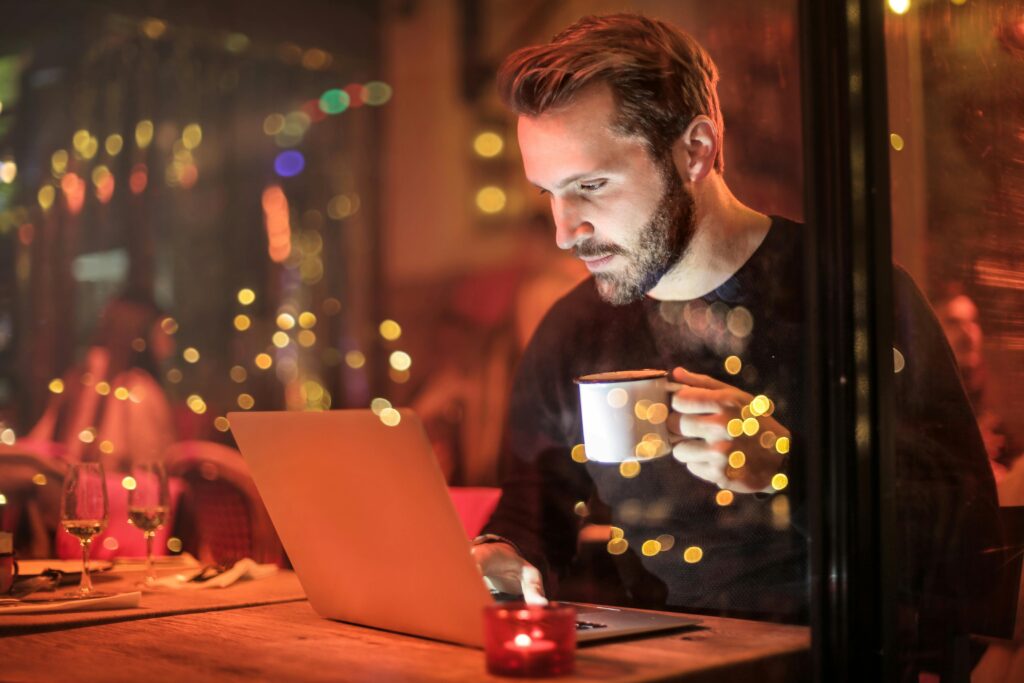 A young man with a beard is working on a laptop on digital marketing at a cozy cafe with warm lighting, holding a coffee cup.